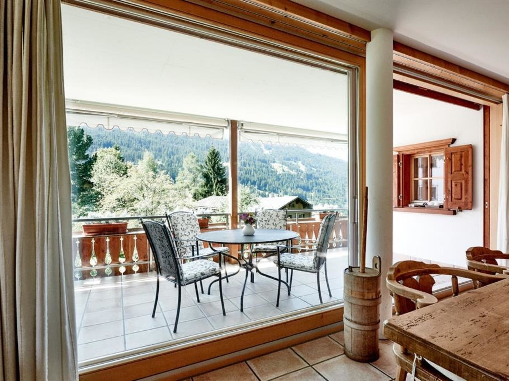 Ein Balkon mit Tisch und Stühlen, einem Holfass und Blick auf ein bewaldetes Gebirge.