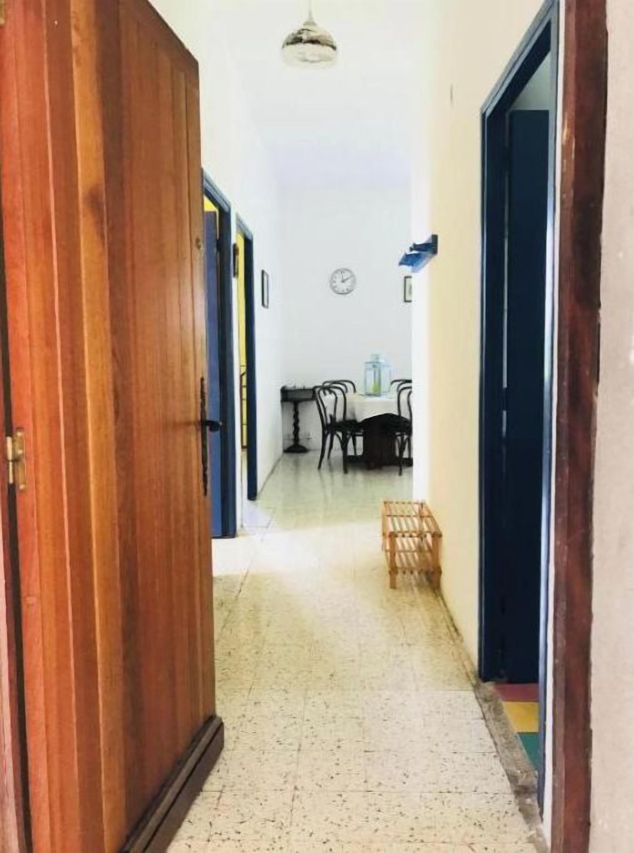 A hallway with a wooden door on the left, a dining area with a table and chairs in the center, and a small table with items on the right.