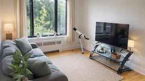 Ein modernes Wohnzimmer mit einer Couch, einem Fernseher auf einem Schrank, einem Teleskop und einem Fenster mit Blick auf Bäume.