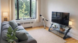 Un soggiorno moderno con un divano, una televisione su un mobile, un telescopio e una finestra che si affaccia su alberi.