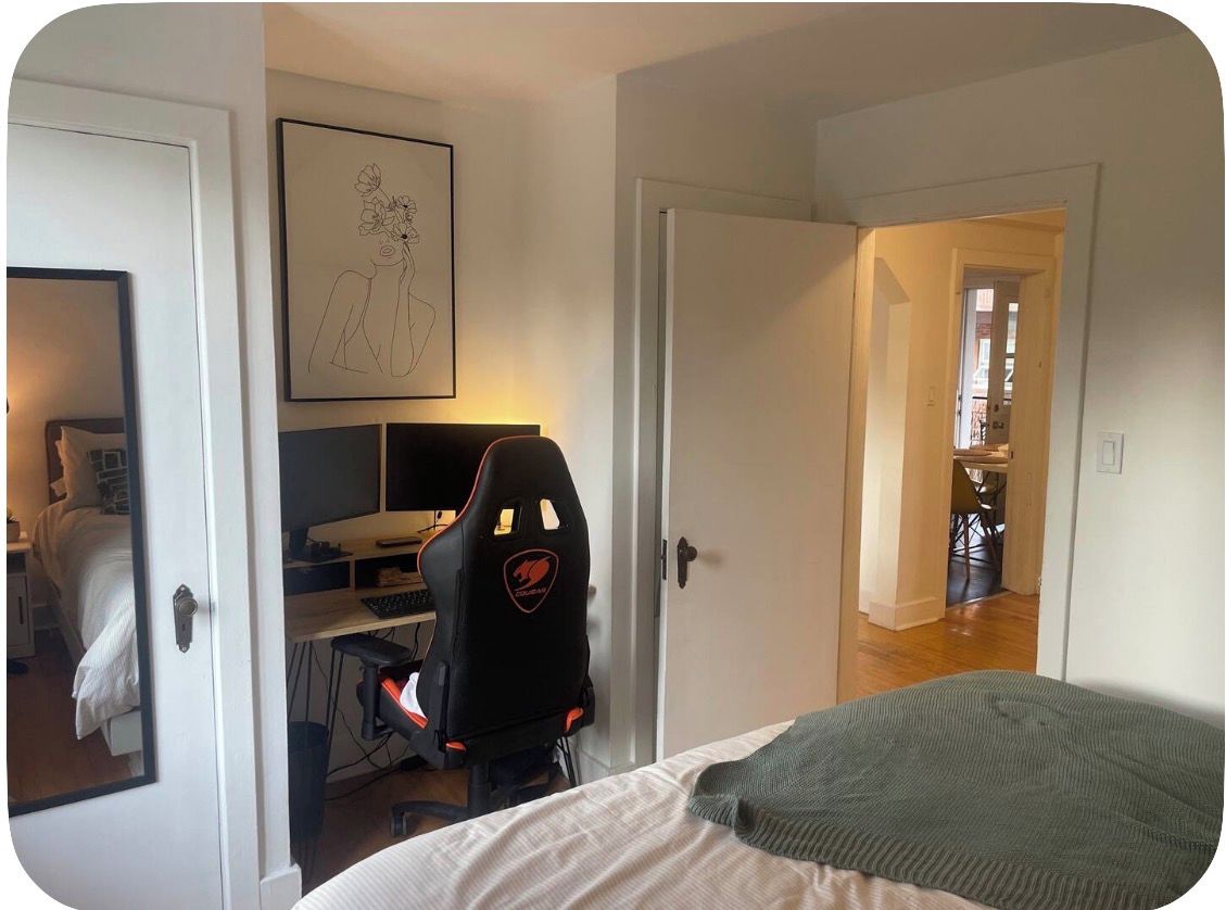 Una camera da letto con un letto, una scrivania con un computer, una sedia e una porta che dà su un'altra stanza.