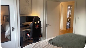 Ein Schlafzimmer mit einem Bett, einem Schreibtisch mit Computer, einem Stuhl und einer Tür zu einem weiteren Raum.