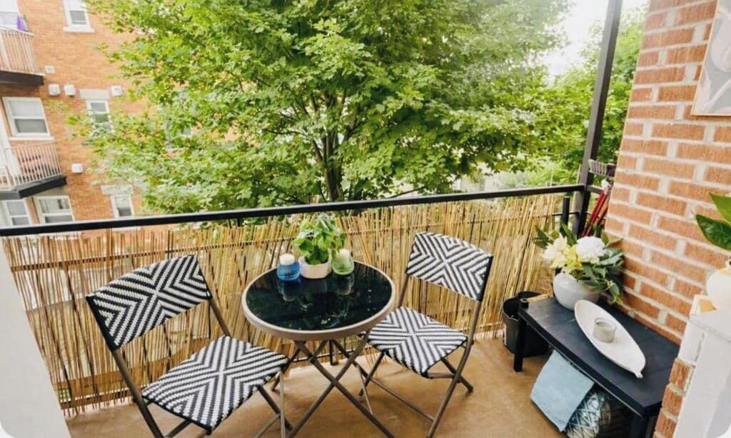 Un balcone con un tavolo rotondo e quattro sedie, un piccolo tavolo nero con un vaso e un piatto, e vista su un albero e degli edifici.