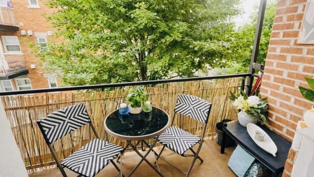 Ein Balkon mit rundem Tisch und vier Stühlen, ein kleiner schwarzer Tisch mit Vase und Teller sowie Blick auf einen Baum und Gebäude.
