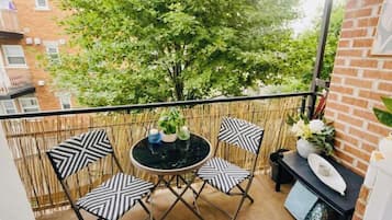 Un balcone con un tavolo rotondo e quattro sedie, un piccolo tavolo nero con un vaso e un piatto, e vista su un albero e degli edifici.