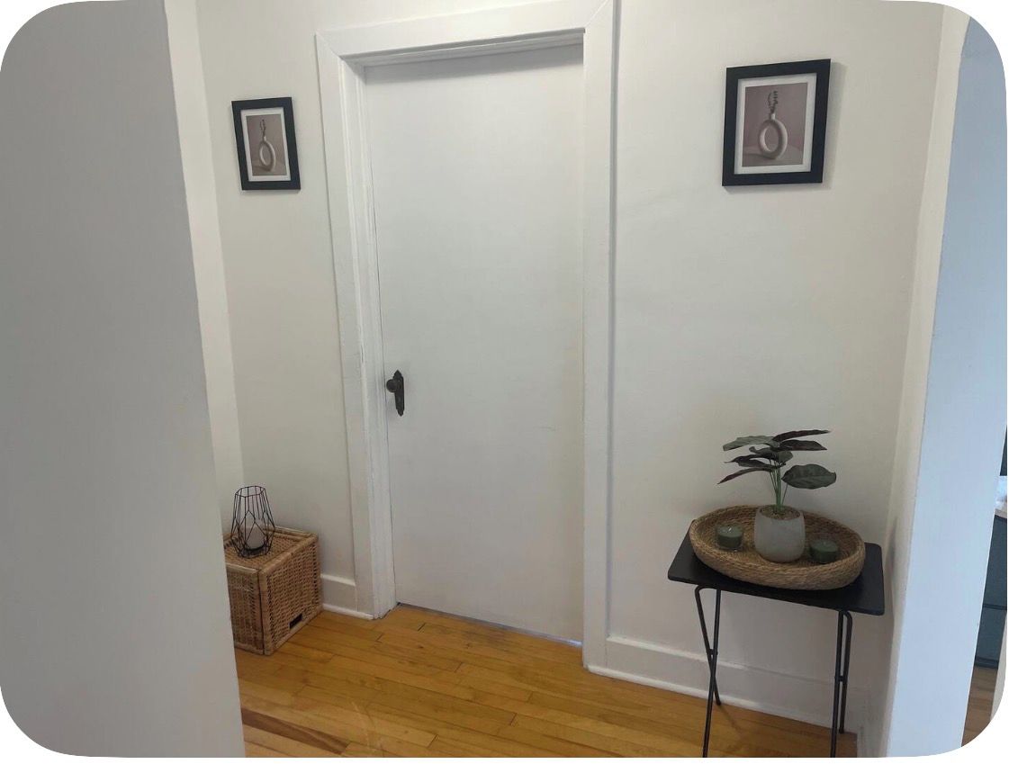 Um corredor com uma porta branca, uma pequena mesa com uma planta e duas quadros nas paredes.