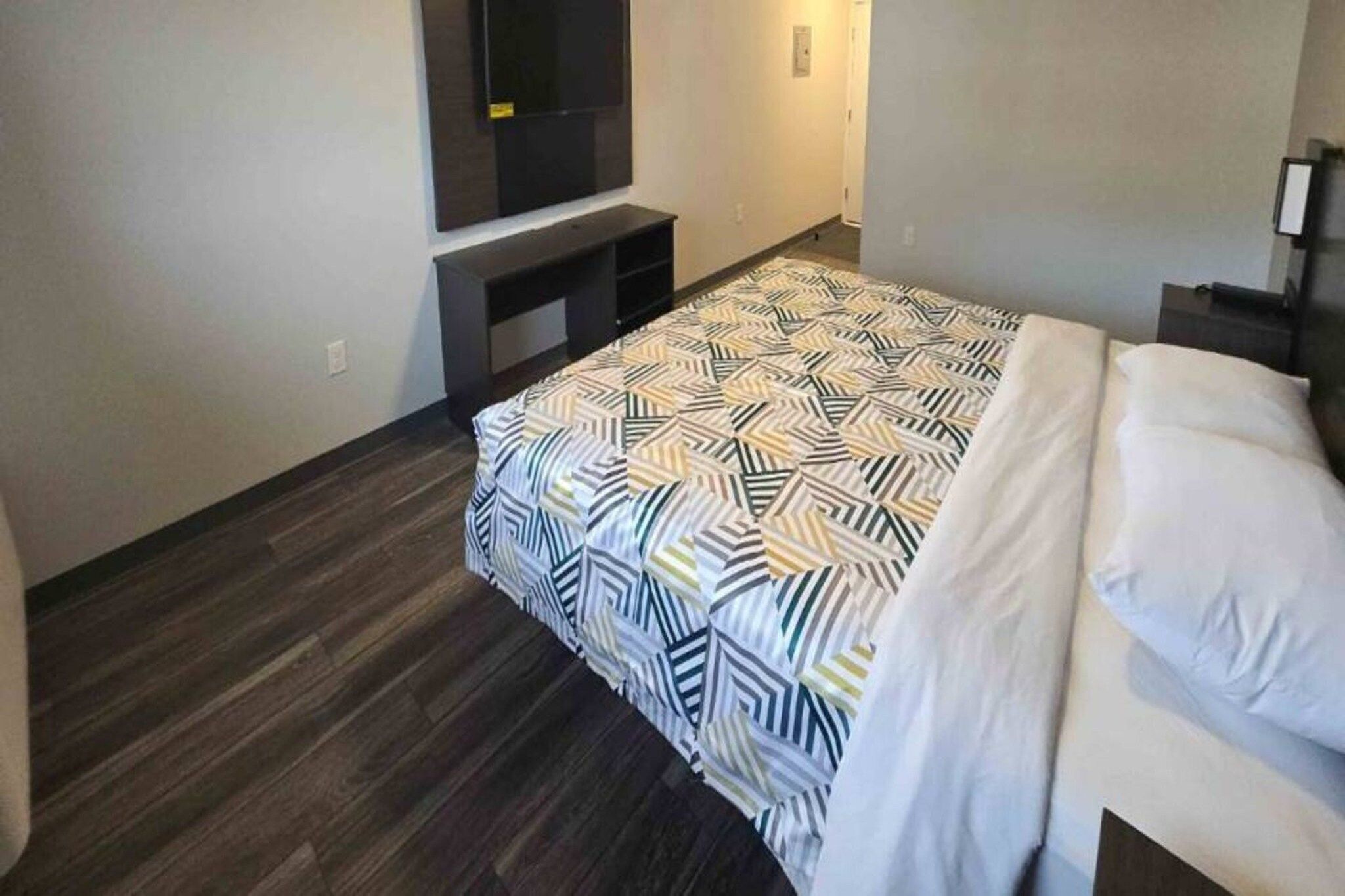 A hotel room with a bed, a TV mounted on the wall, and a dark wooden floor.