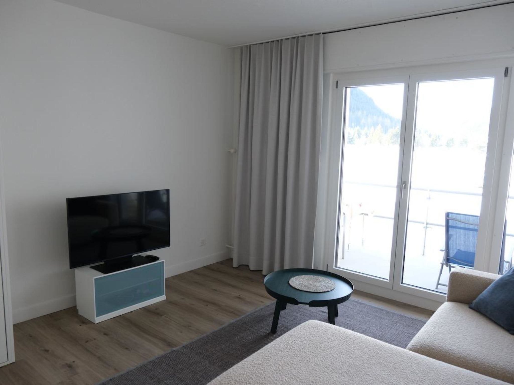 A modern living room with a flat-screen TV, a sofa, a small table, and a view of a balcony with a mountain backdrop.