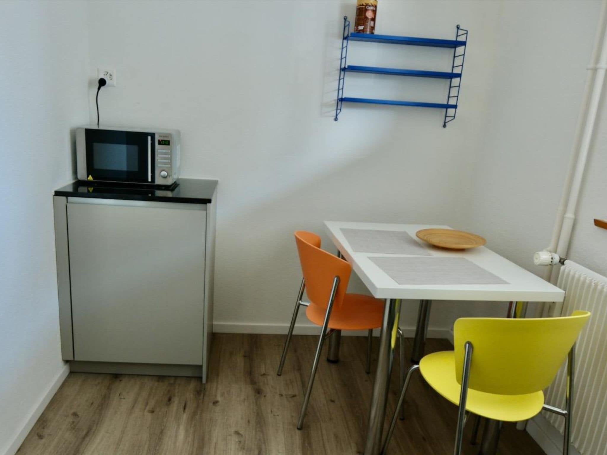 A compact kitchenette with a microwave, a small refrigerator, a white table with a wooden board, and two chairs.