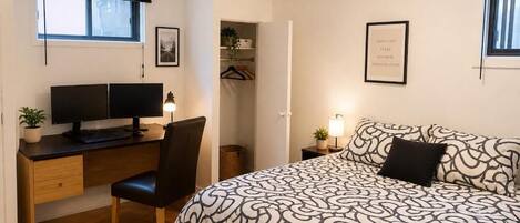 Quarto com cama, escrivaninha, cadeira, televisão e mesa de cabeceira com abajur.
