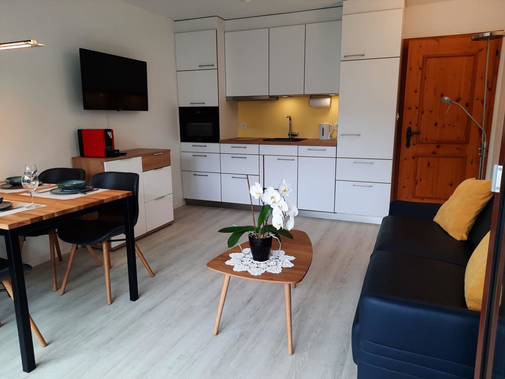 A modern living room with a sofa, a dining table with chairs, a coffee table with a plant, and a kitchen area with cabinets and appliances.