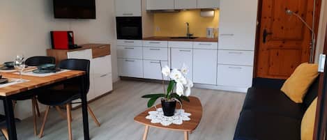 A modern living room with a sofa, a dining table with chairs, a coffee table with a plant, and a kitchen area with cabinets and appliances.