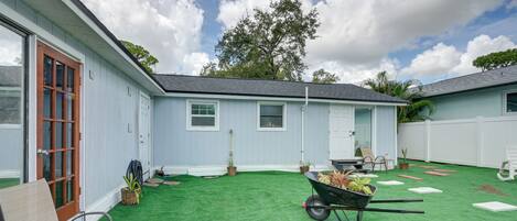 A residential backyard with artificial green turf, a wheelbarrow with plants, a wooden door, and a white fence.