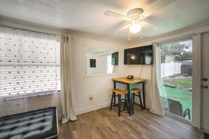 Een kamer met een plafondventilator, een aan de muur bevestigde televisie, een eettafel met twee stoelen en een schuifdeur naar een buitenruimte.