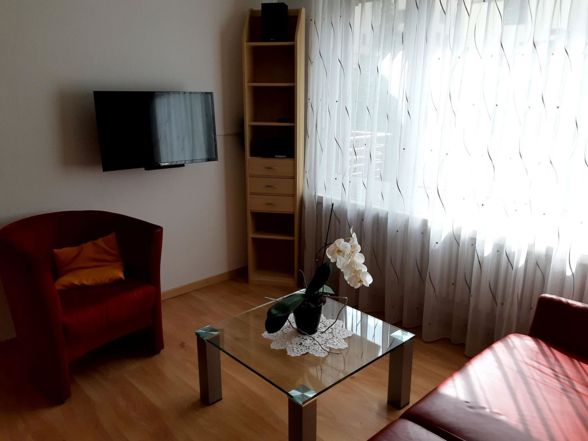 A living room with a red sofa, a glass coffee table with a plant, a TV mounted on the wall, and a wooden bookshelf.