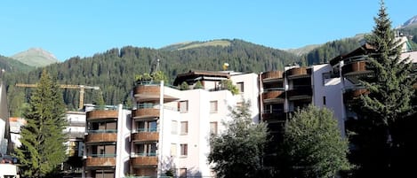 A multi-story building with balconies, surrounded by trees and a mountainous backdrop.