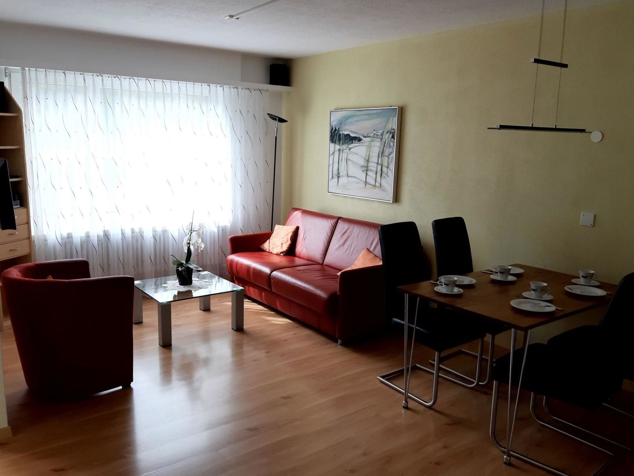 A living room with a red sofa, a glass coffee table, a dining table with chairs, a painting on the wall, and a window with curtains.