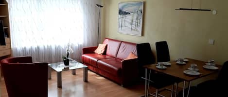 A living room with a red sofa, a glass coffee table, a dining table with chairs, a painting on the wall, and a window with curtains.