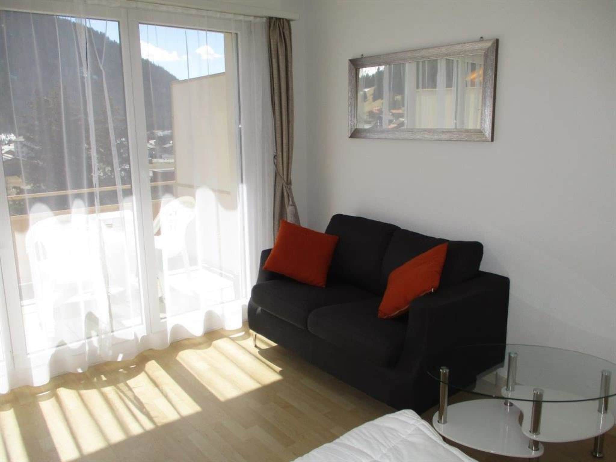 Ein Wohnzimmer mit einer schwarzen Couch, zwei roten Kissen, einem Glastisch und einem großen Fenster mit Vorhängen.