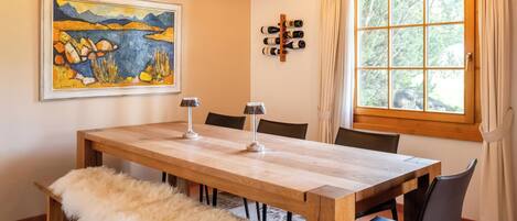 A dining area with a wooden table, chairs, a bench, a painting, a wine rack, and a window with curtains.