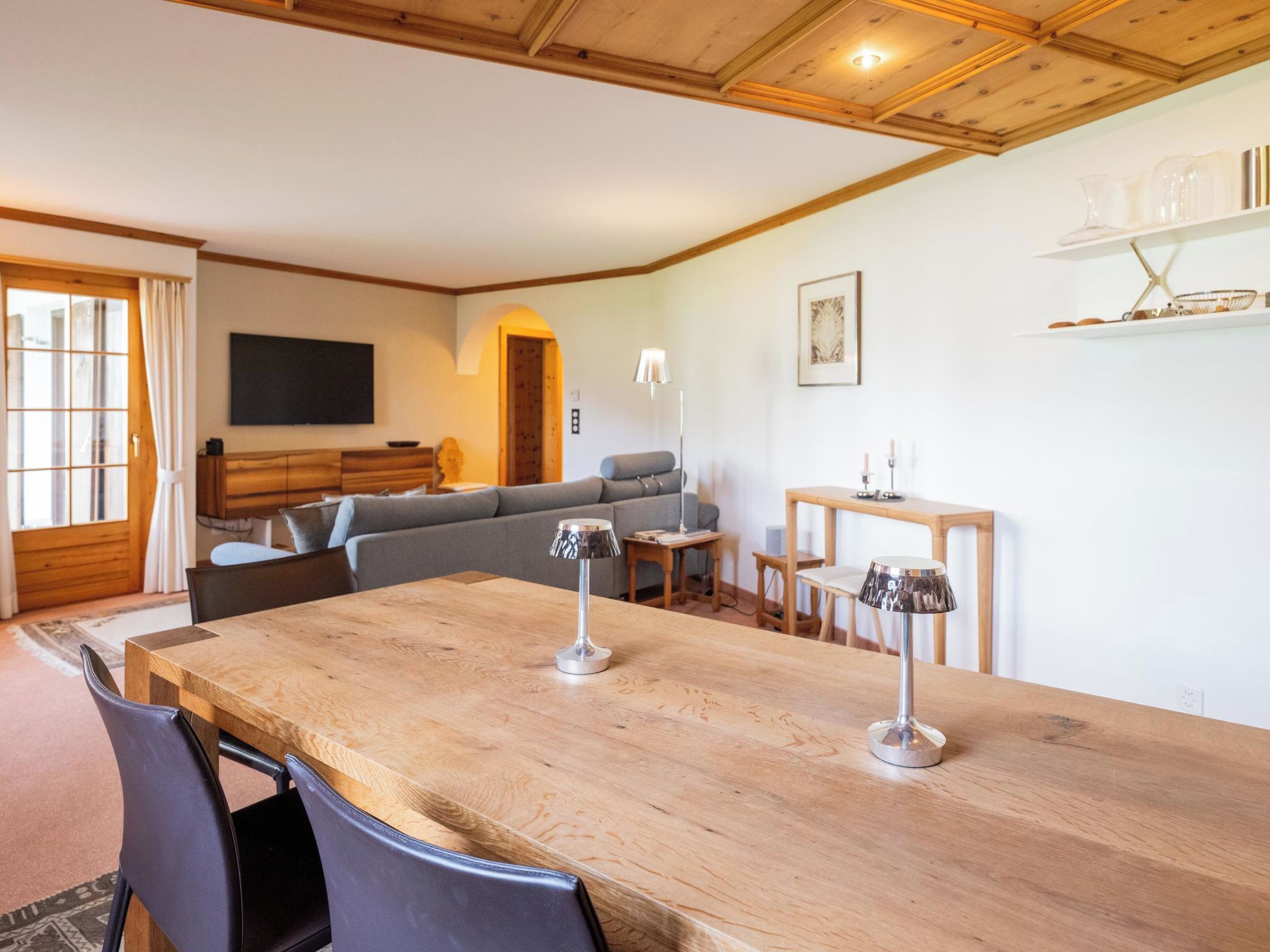 Een ruime woonkamer met een houten eettafel, twee stoelen, een bank en een aan de muur bevestigde televisie.