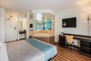 A hotel room with a large bed, a sitting area with a desk and chair, a television, and a bathroom with a bathtub and mirror.