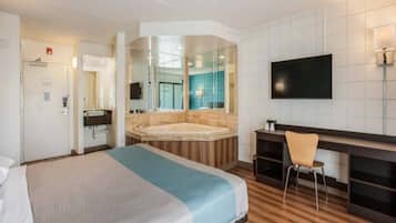A hotel room with a large bed, a sitting area with a desk and chair, a television, and a bathroom with a bathtub and mirror.