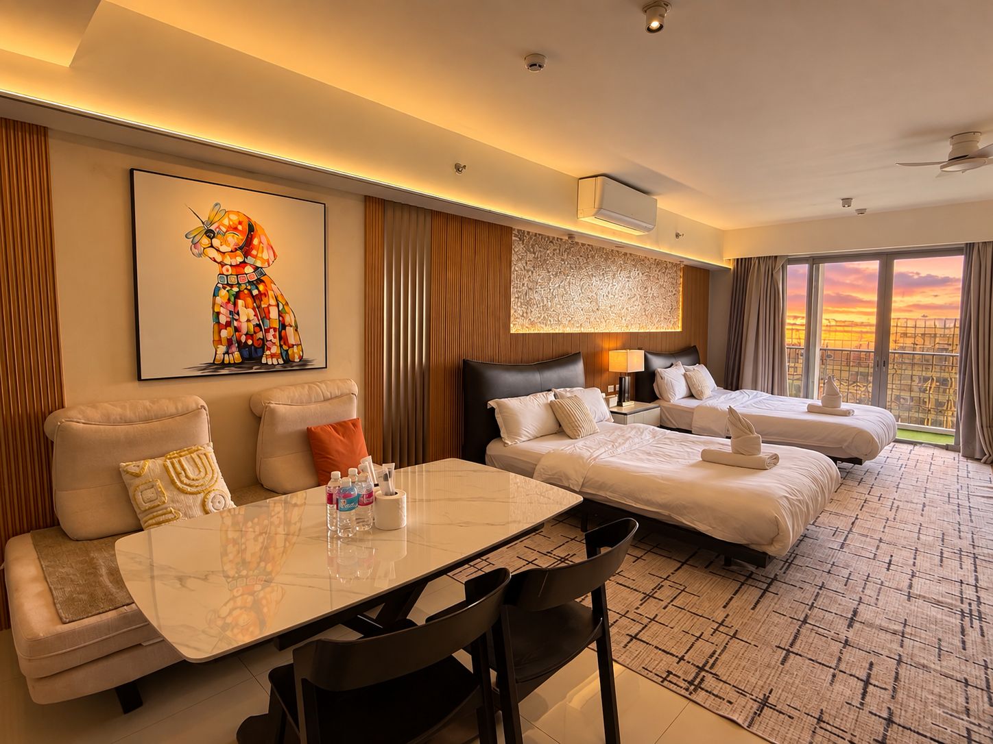 A hotel room with a bed, a sofa, a dining table, and a view of the cityscape.