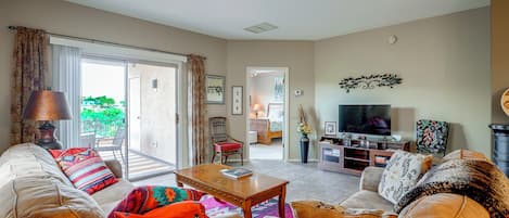 Una sala de estar con sofá, sillón, mesa de centro y televisor. Hay una puerta corrediza de vidrio que da a un balcón.
