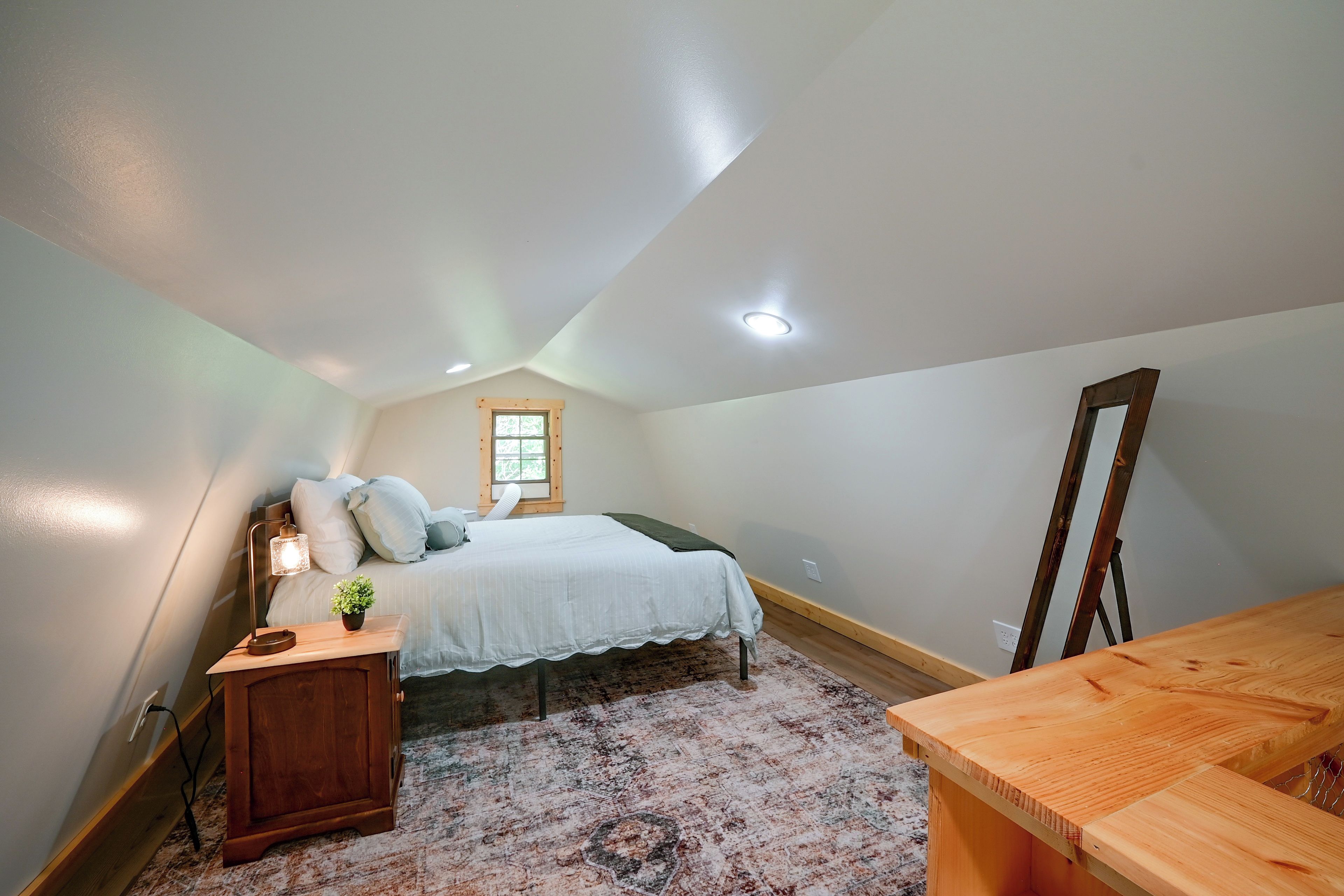 A bedroom with a bed, bedside table, lamp, window, and a mirror.