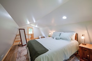 A bedroom with a large bed, a wooden ladder, a small table with a lamp, and a window.
