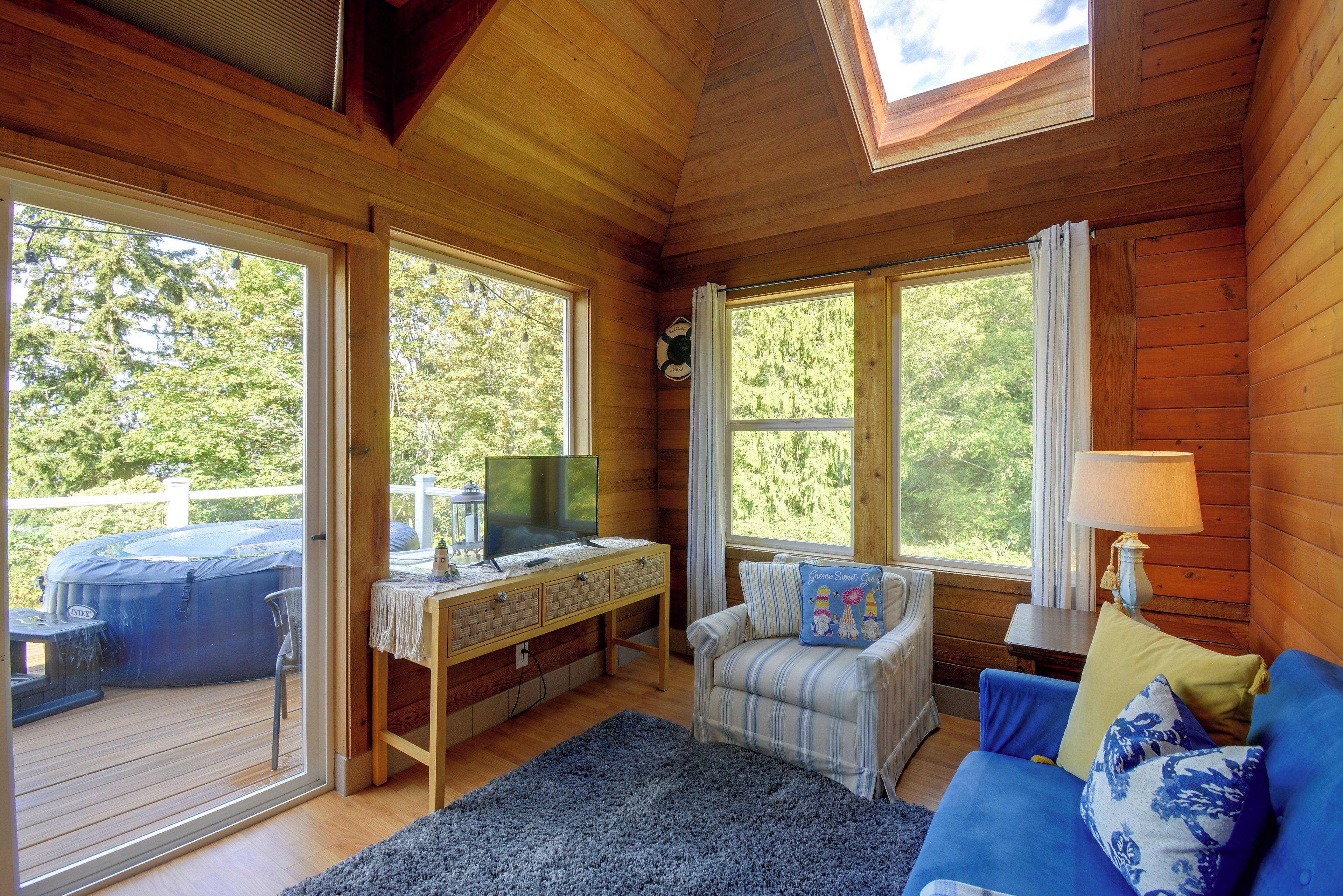 A cozy cabin interior with a wooden ceiling, a sofa, a television on a wooden table, and a view of the outdoors through large windows.