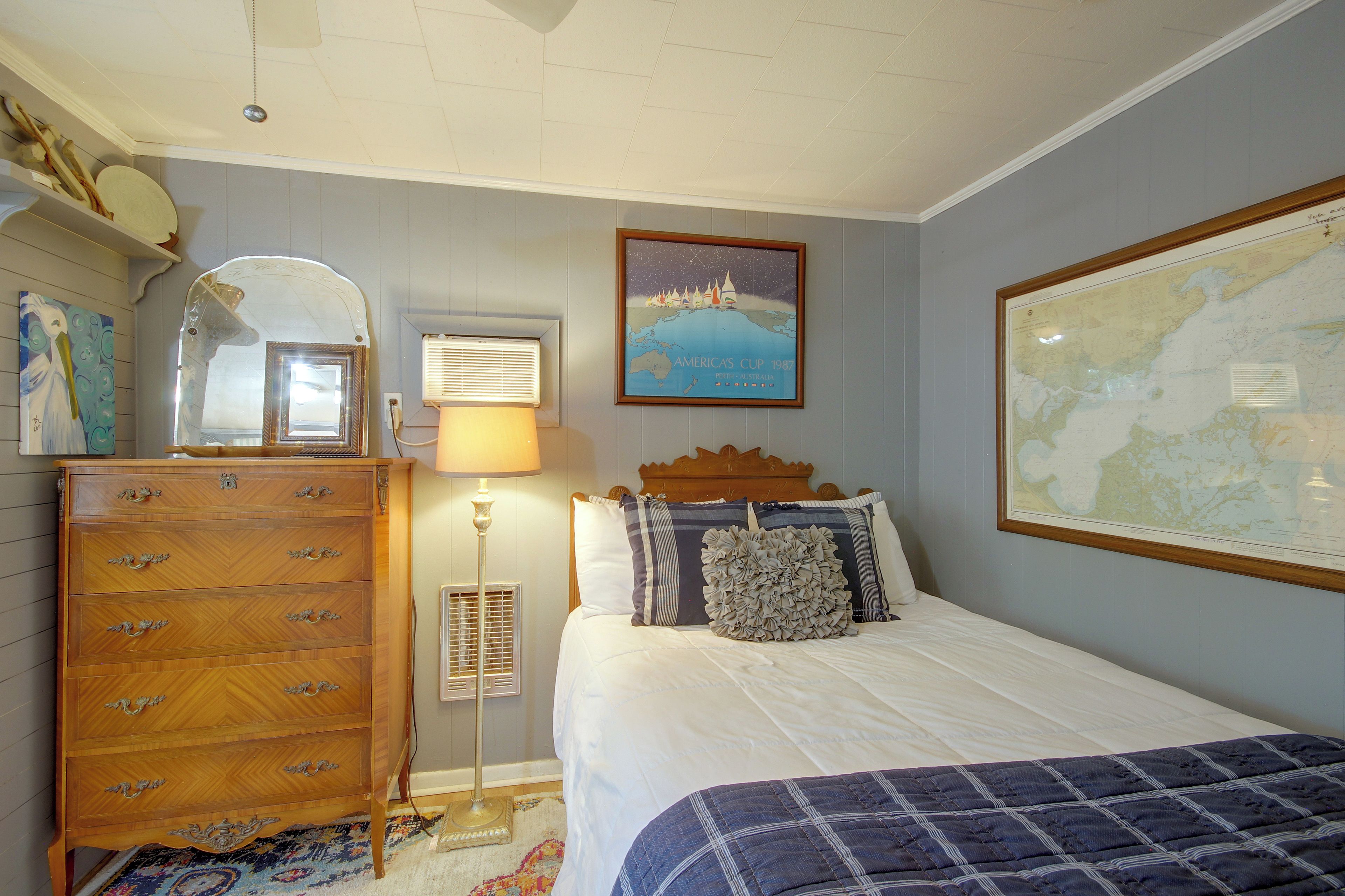 A bedroom with a wooden dresser, a bed with a blue quilt, a map on the wall, and a framed picture of a sailing event.