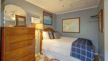 Une chambre avec une commode en bois, un lit recouvert d’une couverture bleue, une carte accrochée au mur et une lampe.