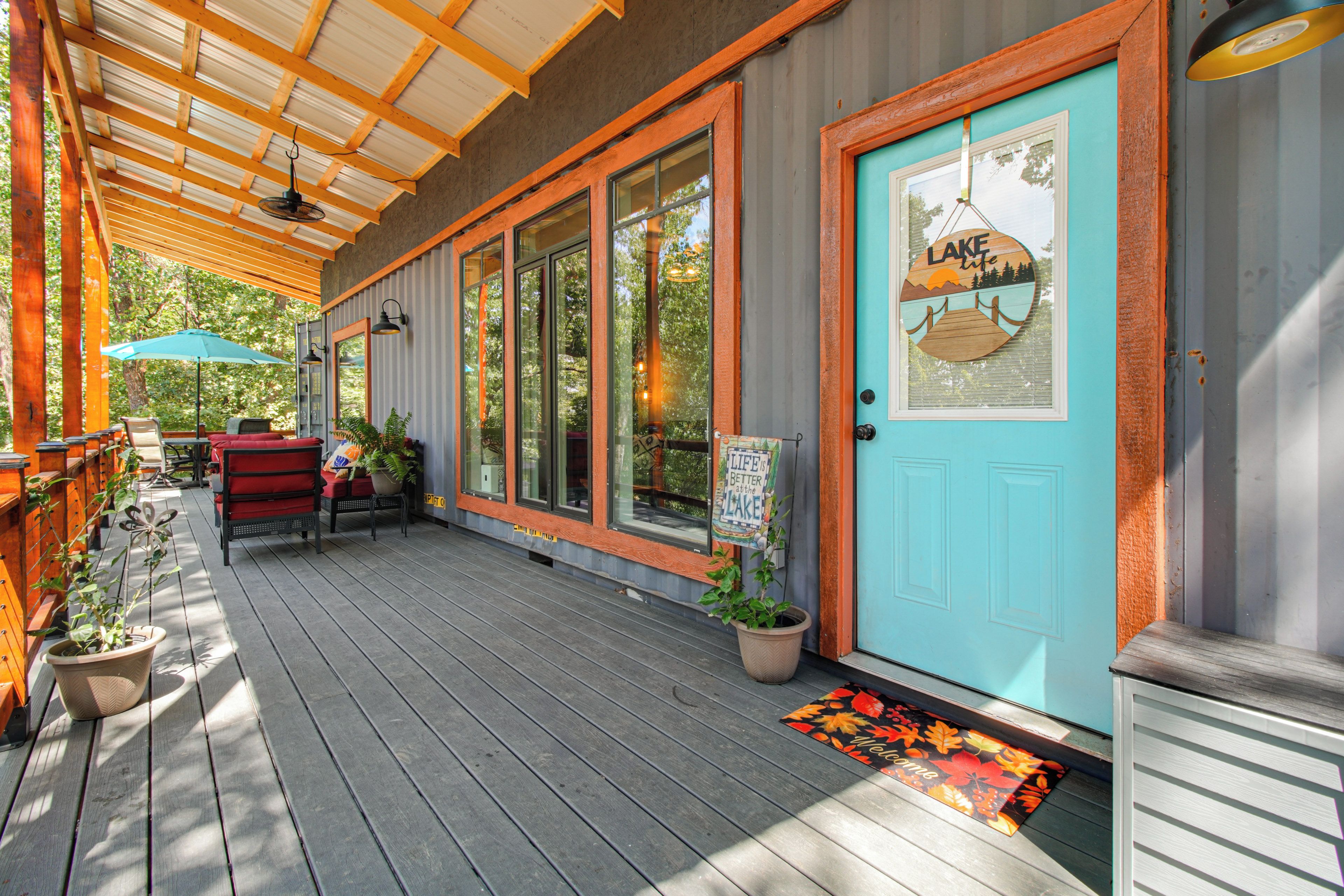 Un porche abrité avec une terrasse en bois, des sièges d’extérieur et une porte ornée d’une pancarte indiquant « Vie au bord du lac ».