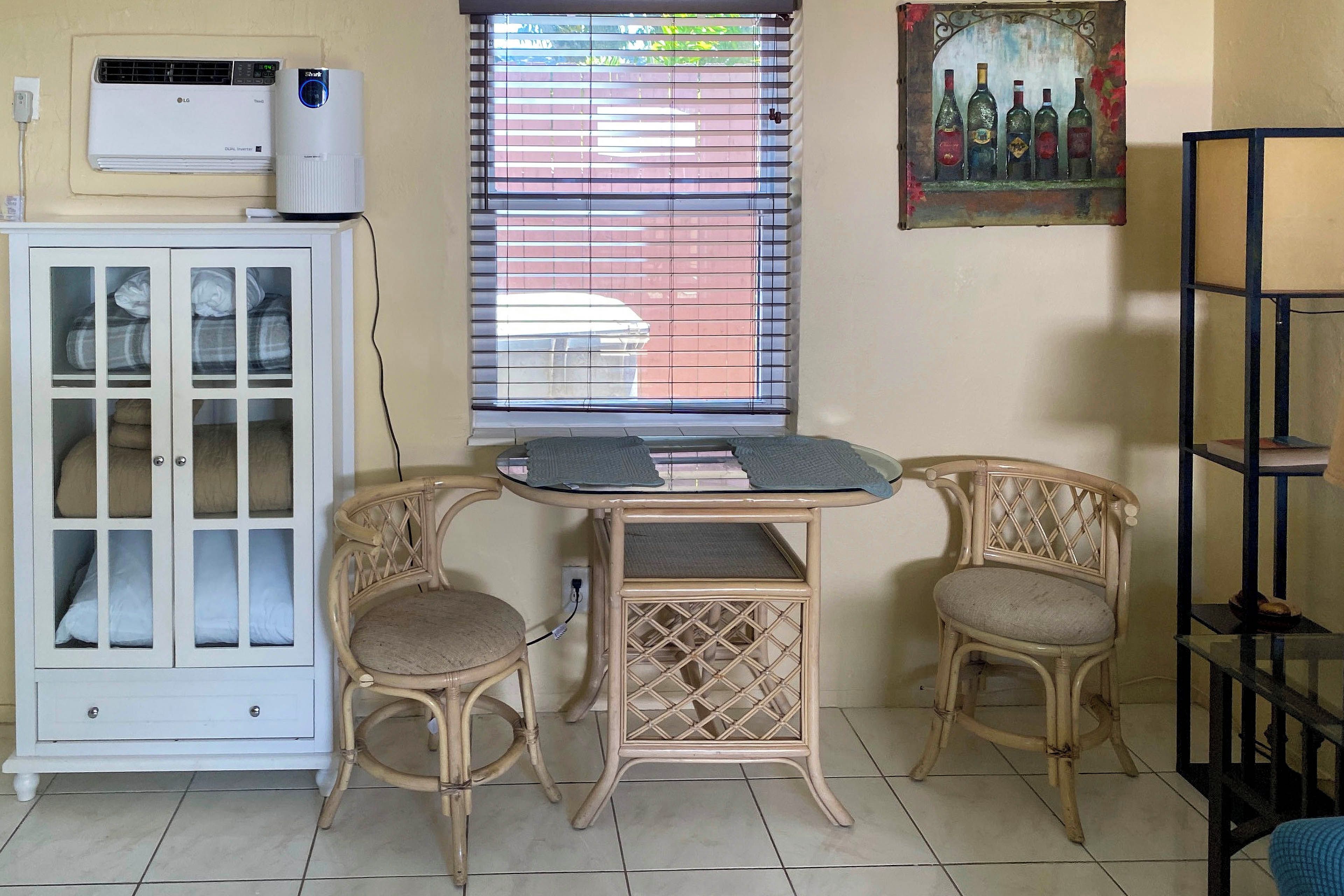 A room with a white cabinet, two wicker chairs, a small table, a window with blinds, and an air conditioner.