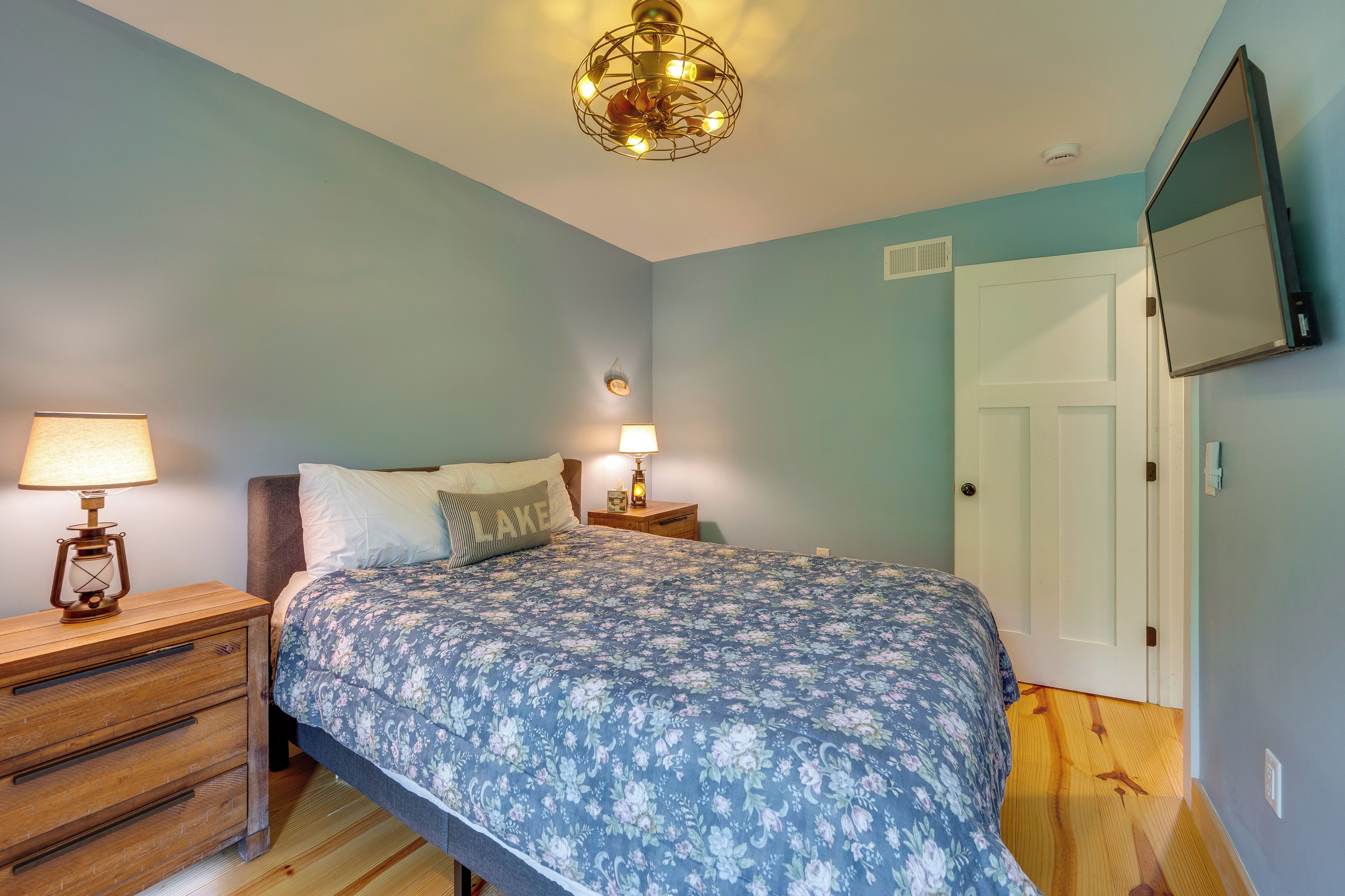 A bedroom with a bed, bedside lamps, a wooden dresser, a wall-mounted TV, and a door.