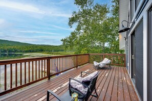 Une terrasse en bois offrant une vue sur un lac et une forêt, deux fauteuils à bascule et une table avec une bouteille.