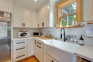 A modern kitchen with white cabinetry, a stainless steel refrigerator, a microwave, a sink with a black faucet, and a window with a view of trees. - On Neal Pond! Spacious Group Retreat w/ Game Room (Lunenburg)