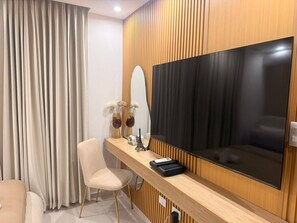 A modern hotel room with a large flat-screen TV mounted on a wooden panel wall, a comfortable chair, a small table with decorative items, and a large window with curtains. - Nice apartment close to everything (Santo Domingo Norte)