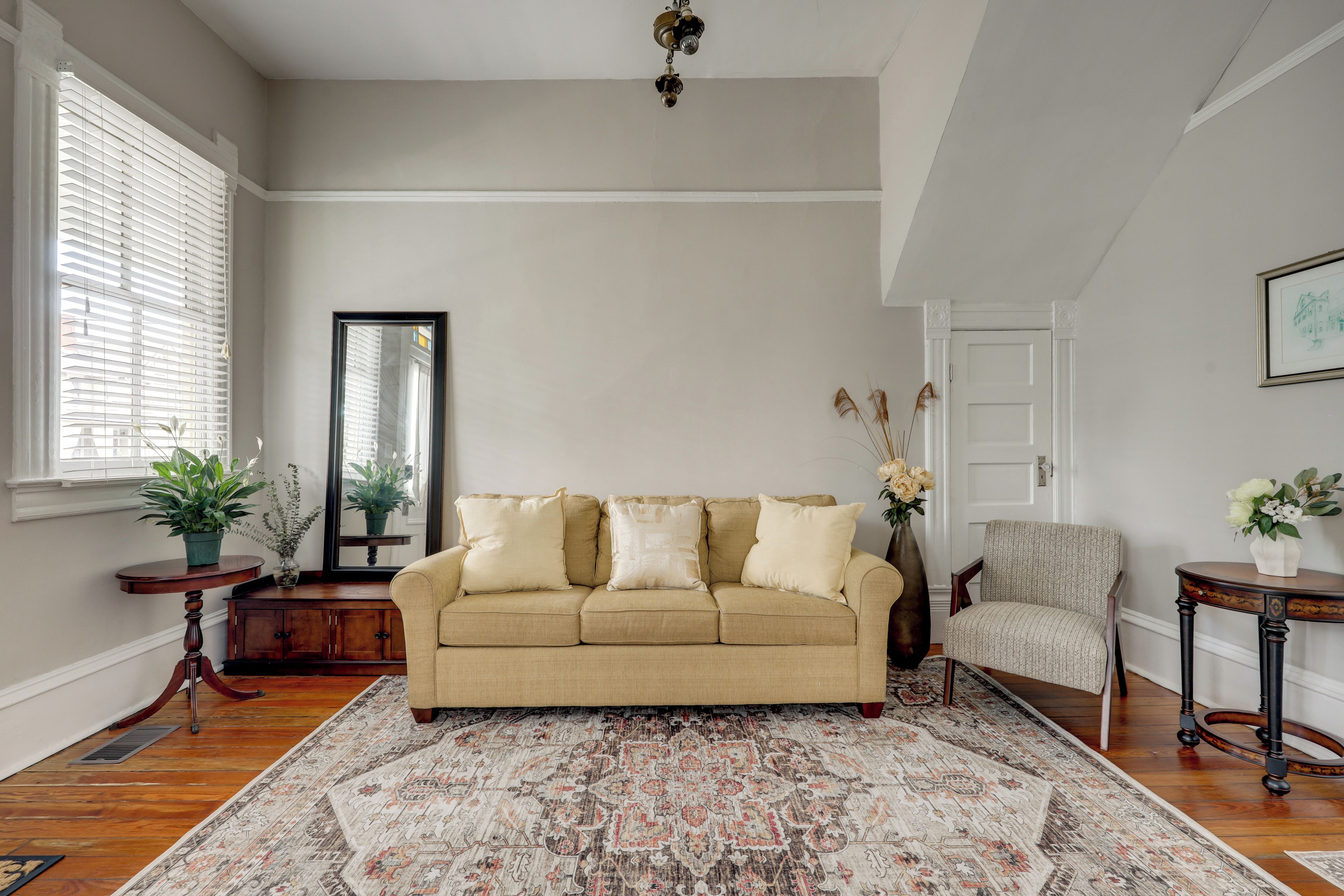 Een woonkamer met een beige bank, een kamerkleed met dessin, een houten vloer, een spiegel, een deur en een kleine tafel met een vaas.