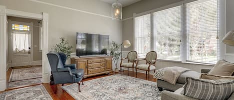Un salon doté d’un grand téléviseur à écran plat, d’un fauteuil en cuir bleu, d’une commode en bois, de deux fauteuils rembourrés, d’un tapis à motifs et de grandes fenêtres équipées de stores.