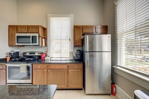 A kitchen with wooden cabinets, a stainless steel refrigerator, a microwave, a stove, and a window with blinds. - Historic Jacksonville Home: Near EverBank Stadium (Jacksonville)