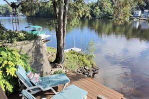 A wooden deck with two lounge chairs and a small table, overlooking a calm lake with a dock and a birdhouse. - Pine Point: 3BR, 2BA, Private Dock & Kayaks! (Holland)
