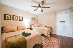 A hotel room with two beds, a ceiling fan, a window with an air conditioning unit, and framed pictures on the wall.