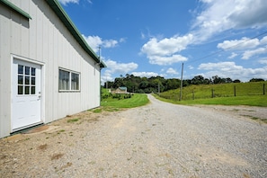A gravel driveway leading to a white building with a green roof, a clear sky, and a green field in the background.