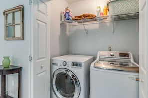 A laundry room with a washing machine, dryer, and shelves above containing cleaning supplies. - Pet-Friendly Private Yard Near Charleston, Beaches (Goose Creek)