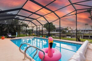 Une piscine agrémentée d’un flotteur en forme de flamant rose, entourée de chaises longues et d’un espace abrité offrant une vue sur les palmiers et les bâtiments.