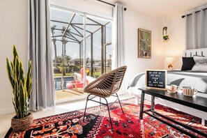 A modern hotel room with a large sliding glass door leading to a pool area, a wicker chair, a bed with a black and white striped bedspread, and a sign with a message.