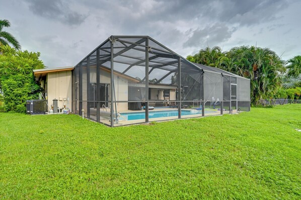 A covered outdoor swimming pool with a glass enclosure, surrounded by a well-maintained lawn and tropical vegetation.